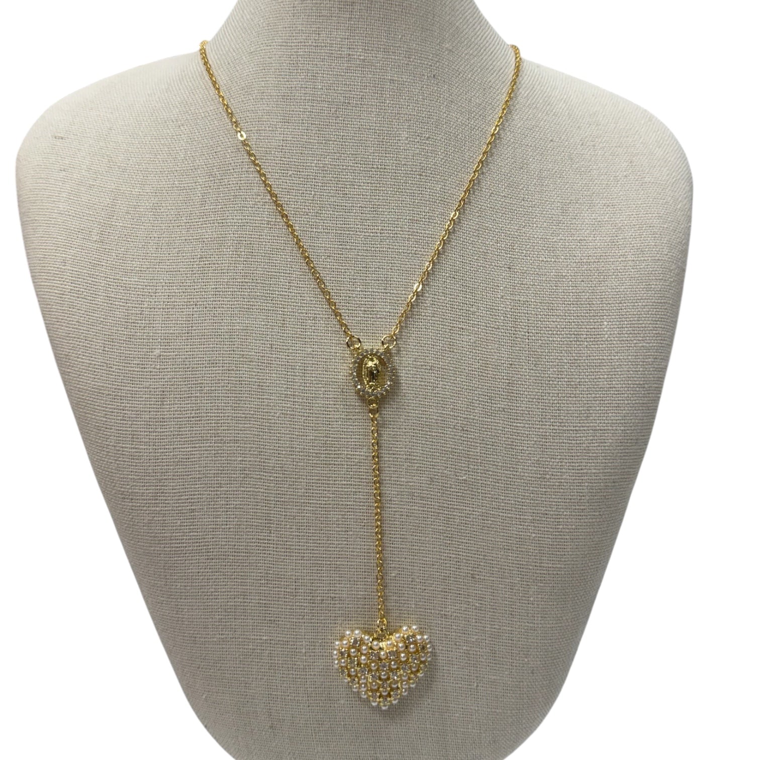 A gold lariat-style necklace displayed on a beige fabric-covered jewelry bust. The necklace features a delicate chain, a small oval medallion, and a long drop ending in a heart-shaped cluster of clear rhinestones.