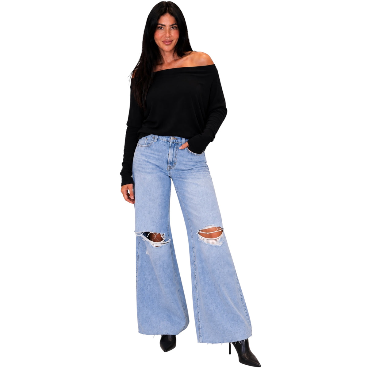 A woman modeling light-wash wide-leg distressed jeans with ripped knees, paired with a black off-the-shoulder long-sleeve top and black pointed boots, against a plain white background.