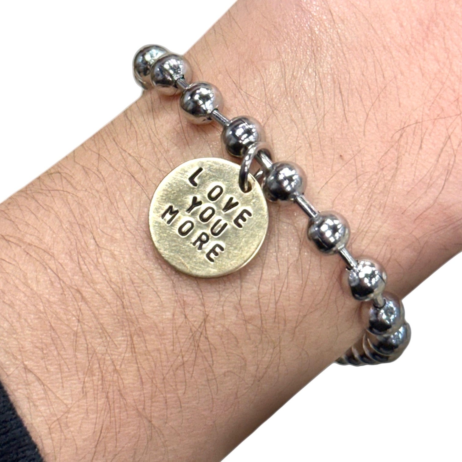Silver beaded bracelet with a gold tag reading 'LOVE YOU MORE' on a wrist.