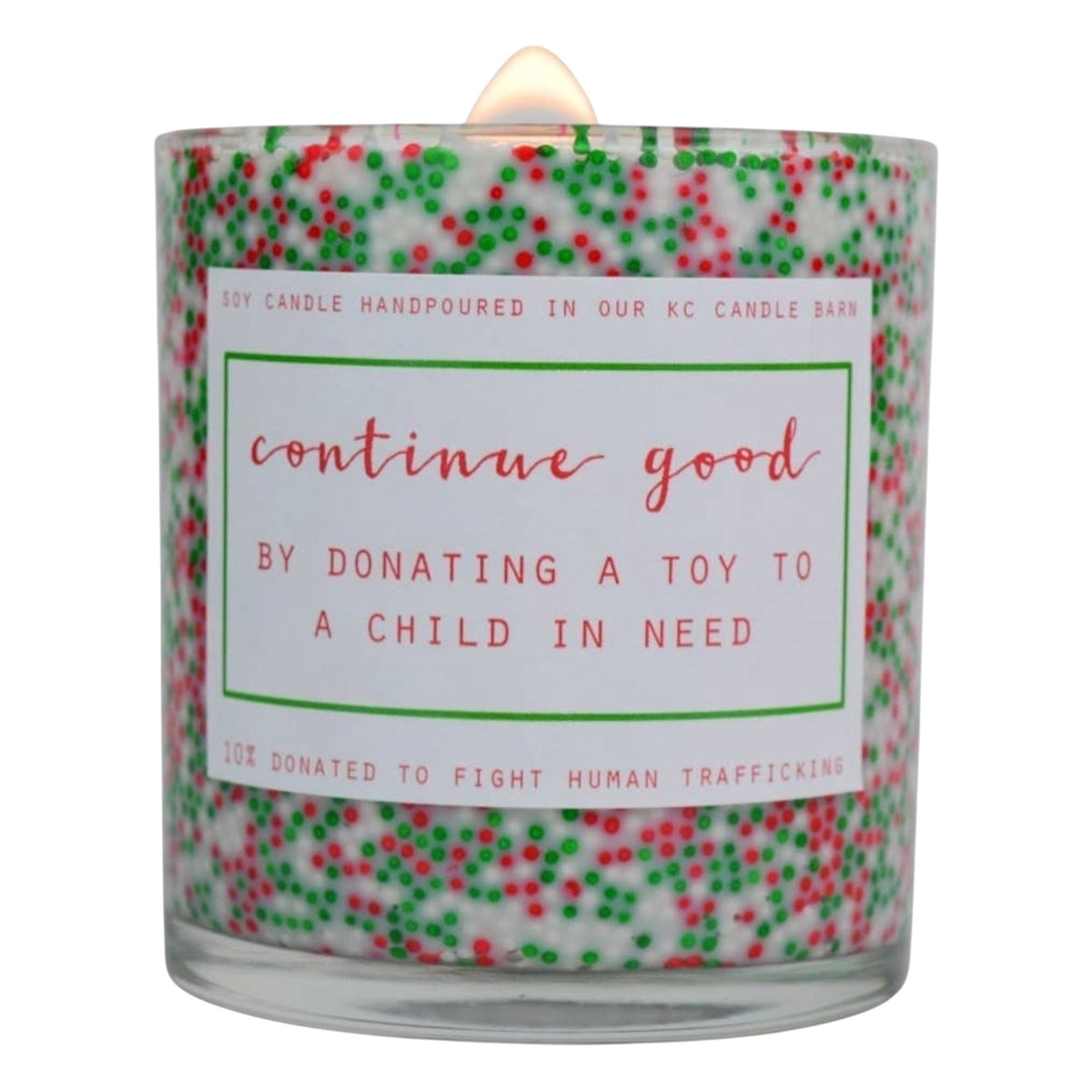 A glass jar candle with a white wax body filled with red and green sprinkles, featuring a white label with red text that reads 'continue good BY DONATING A TOY TO A CHILD IN NEED'.