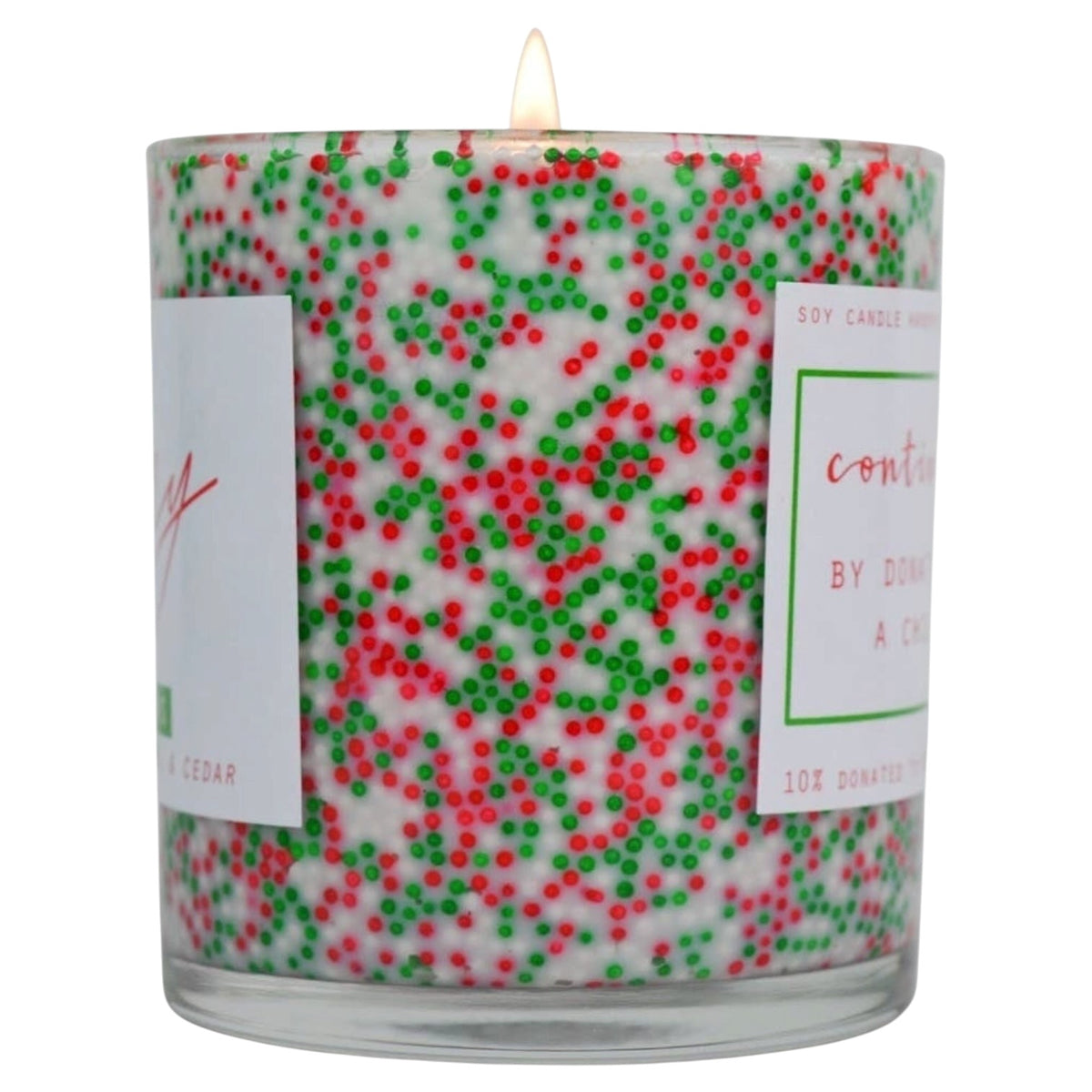 A lit soy candle in a clear glass jar filled with white wax and decorated with red and green sprinkles, with white paper labels on the sides.