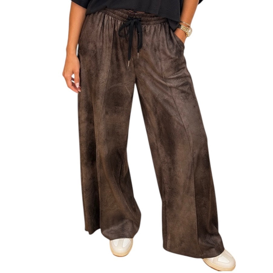 A person wearing wide-leg brown faux-leather pants with a black drawstring waist, paired with a black top and white sneakers, shown against a plain white background.