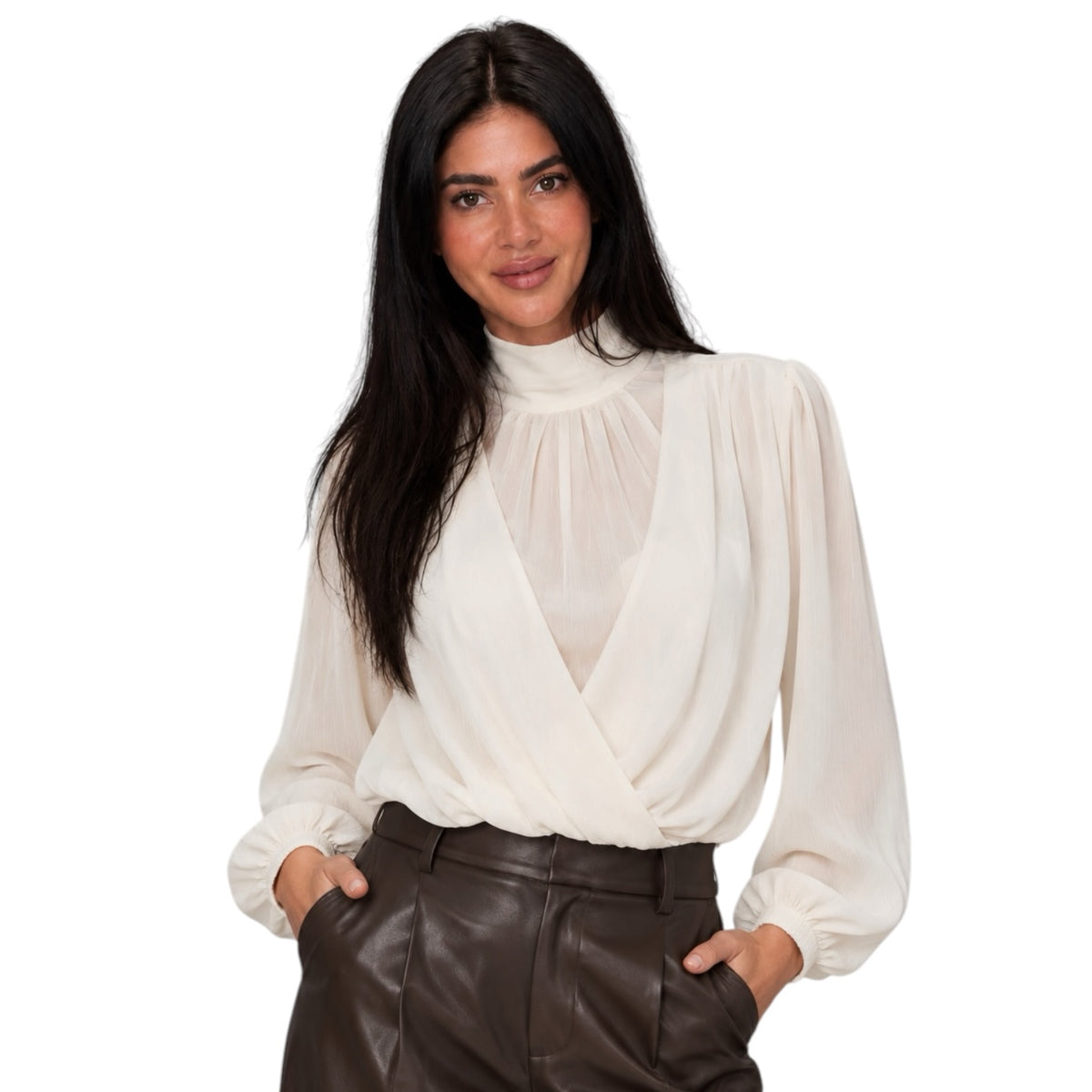 A woman modeling a cream-colored long-sleeve blouse with a high mock neck and gathered front, paired with dark brown faux-leather trousers, against a solid white background.