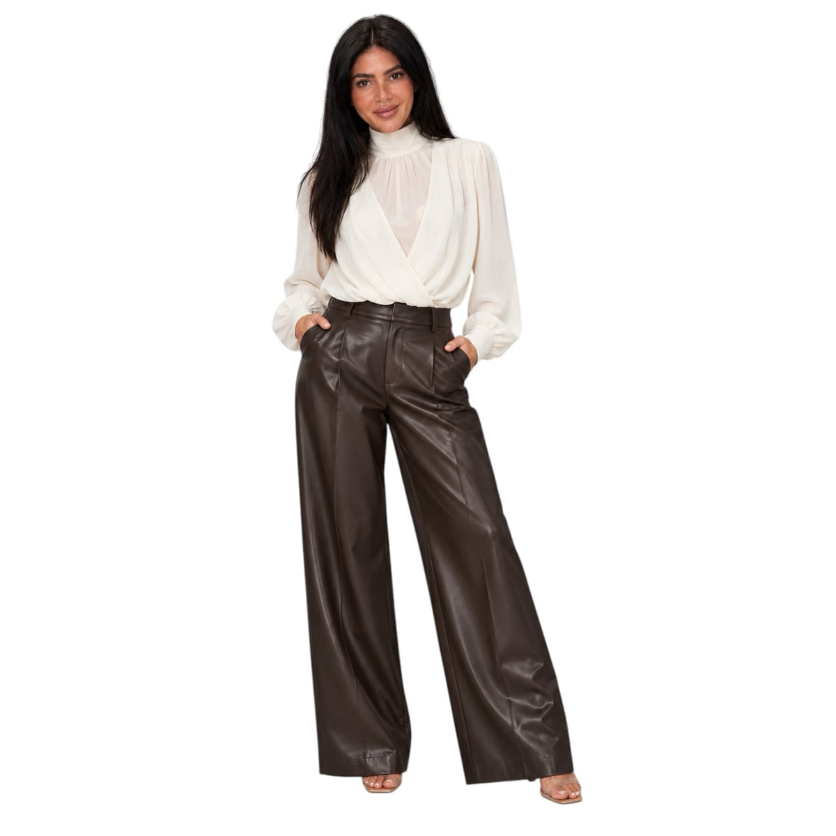 A woman modeling a white long-sleeve mock neck blouse and high-waisted, wide-leg dark brown faux-leather pants against a plain white background.