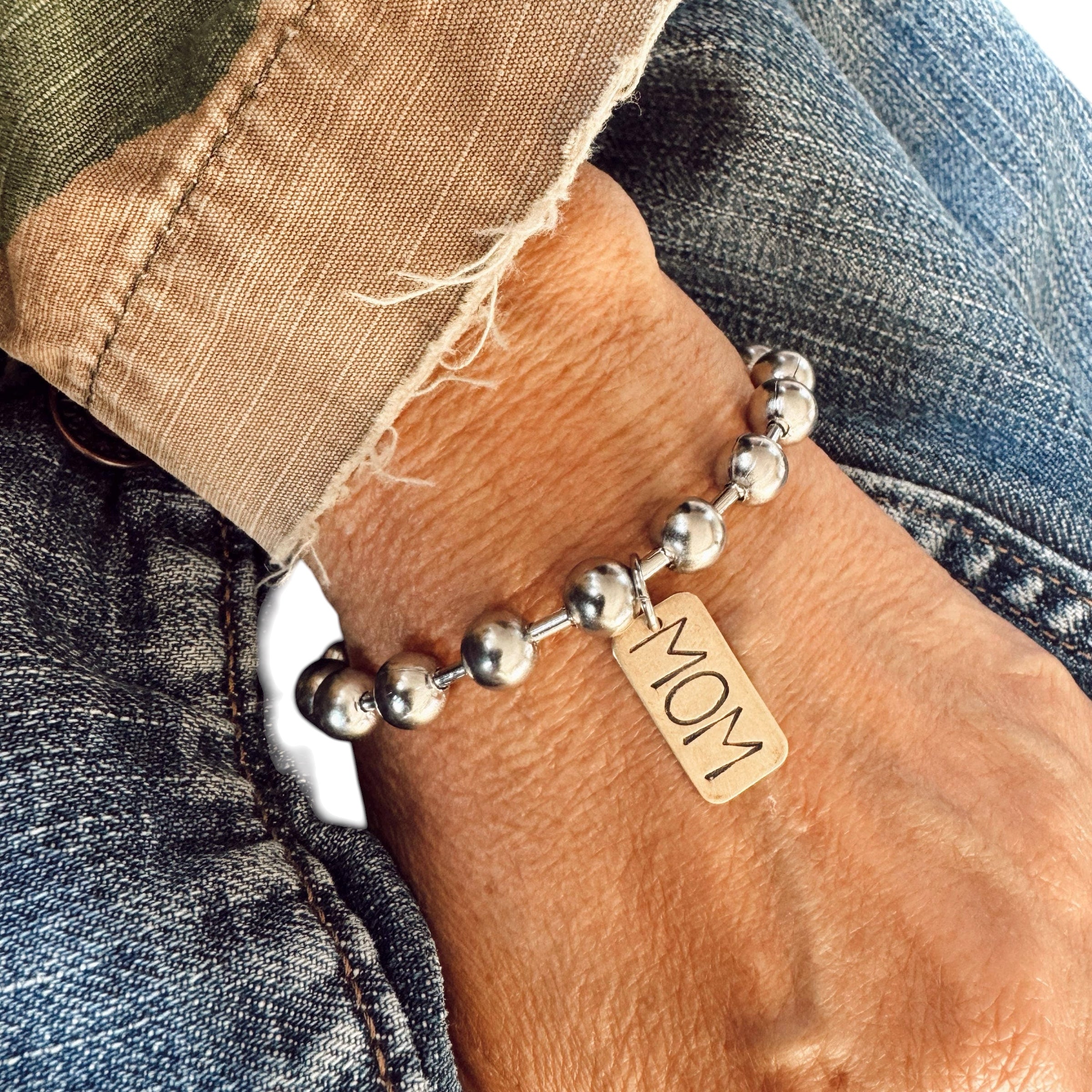 Silver beaded bracelet with a 'MOM' tag on a wrist, wearing jeans.