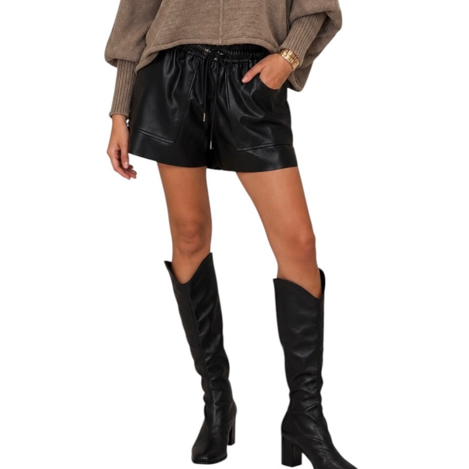 A model wearing black faux-leather drawstring shorts with side pockets, styled with a brown knit sweater and black knee-high boots against a white background.