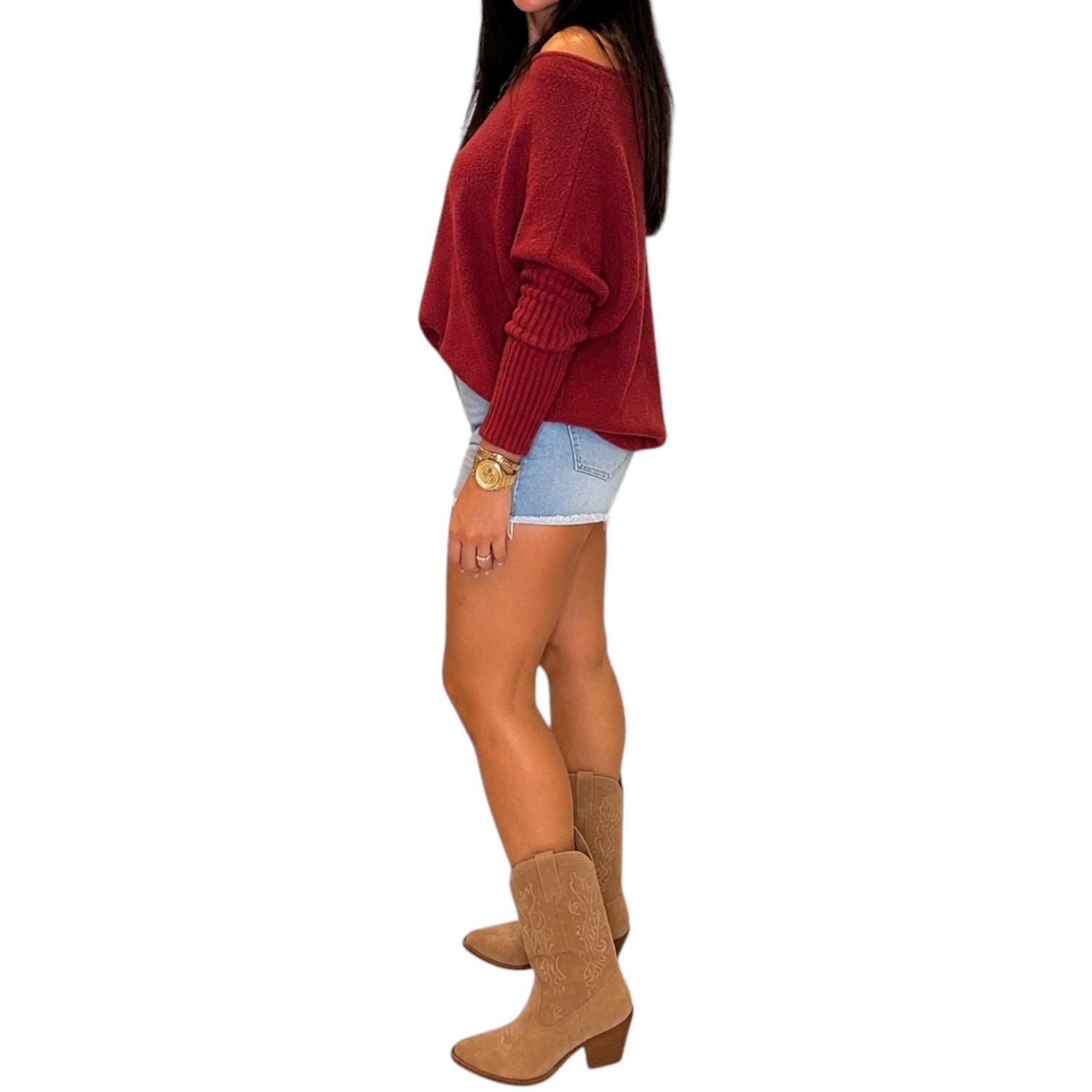 A woman shown from the side wearing a loose off-the-shoulder knit sweater in burgundy, light denim shorts, and tan ankle boots, against a plain white background.