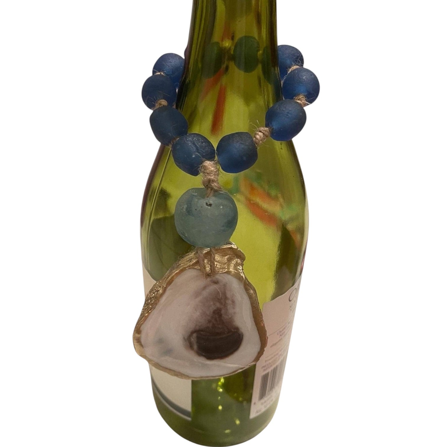 A handmade beaded necklace with blue and light blue beads, featuring a large natural oyster shell pendant with a gold-toned cap, displayed wrapped around a green glass bottle against a white background.