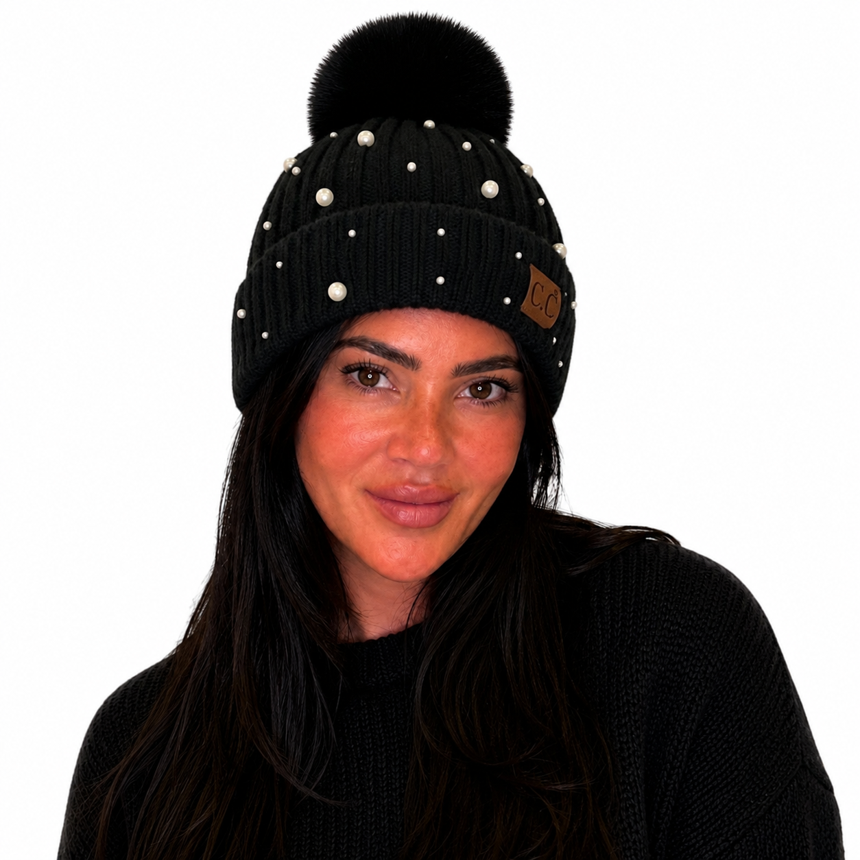 A woman wearing a black ribbed knit beanie with a large black pom-pom, white pearl-like studs, and a small brown leather patch on the side, shown against a plain white background.