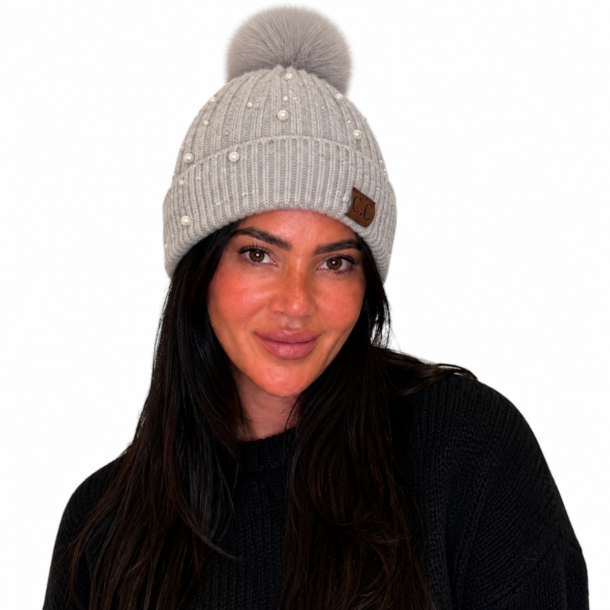 A woman wearing a light gray ribbed knit beanie with a large gray pom-pom, pearl embellishments, and a small brown leather patch on the side, set against a plain white background.
