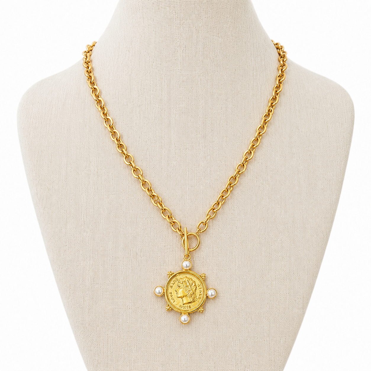 A gold chain necklace with a circular coin pendant and pearl-like accents, displayed on a cream-colored jewelry bust against a white background.
