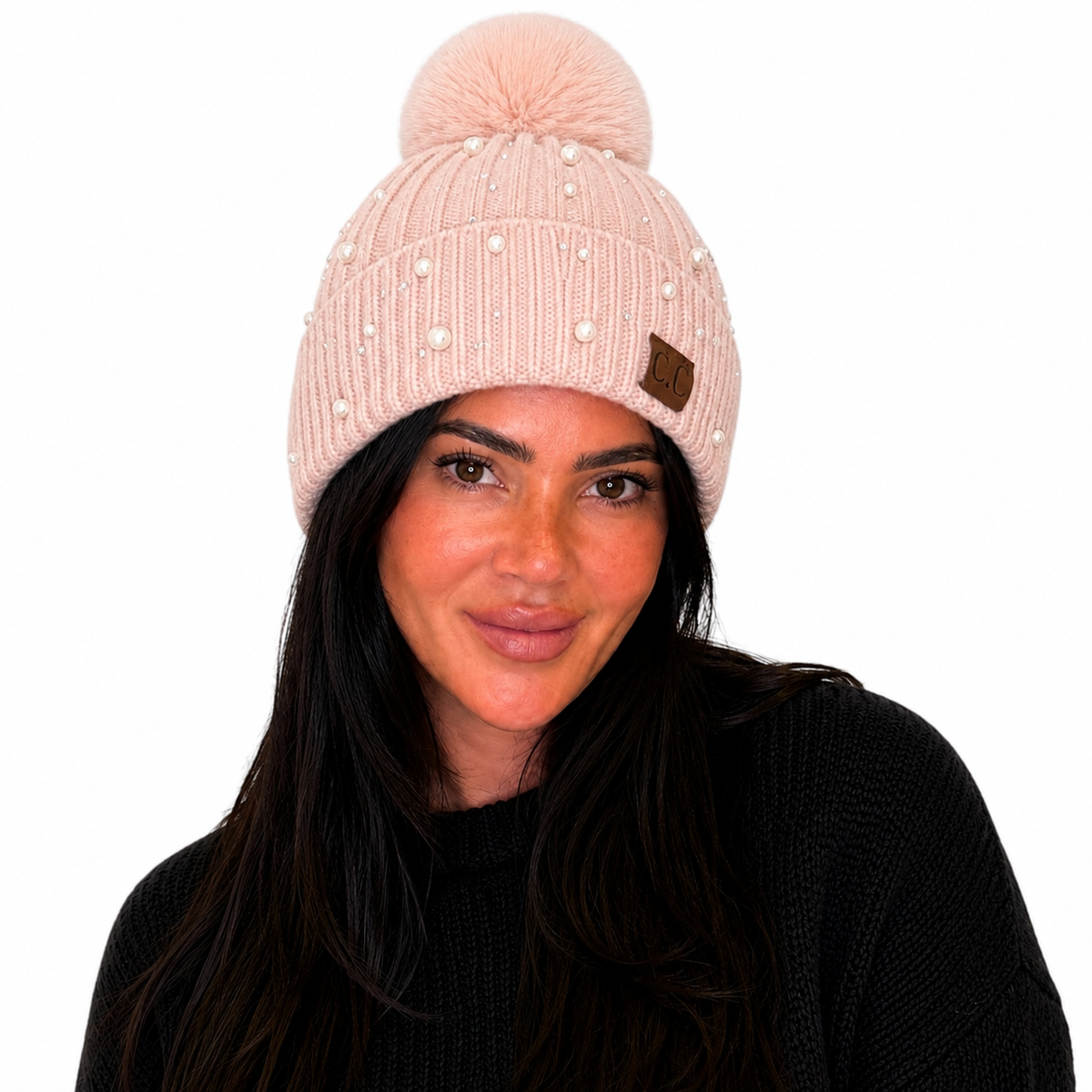A woman wearing a light pink ribbed knit beanie with a matching pom-pom, decorated with small pearl-like embellishments and a small brown leather patch.