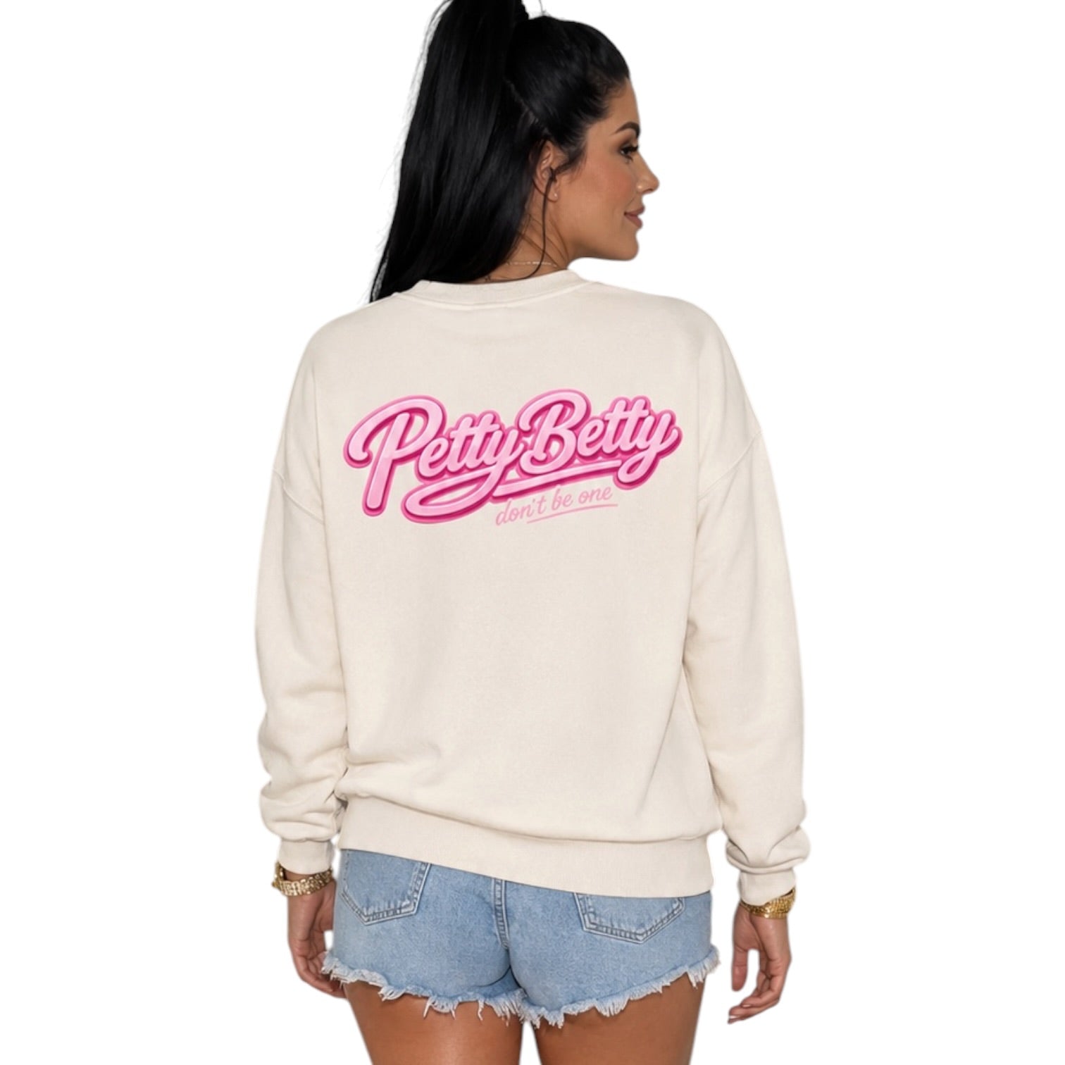 A woman shown from the back wearing a cream-colored crewneck sweatshirt with a pink 'Petty Betty' graphic on the back, paired with light blue denim shorts, against a plain white background.