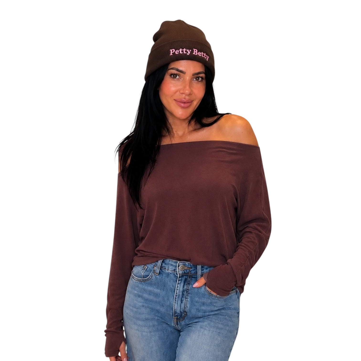 A woman modeling a dark brown off-the-shoulder long-sleeve top and a matching dark brown knit beanie with 'Petty Betty' embroidered in pink on the front, paired with blue jeans against a plain white background.