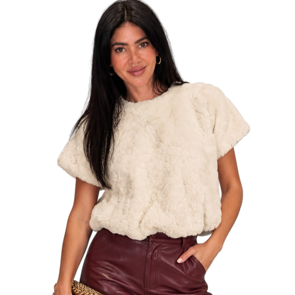 A woman modeling a short-sleeve cream faux fur top paired with burgundy faux leather pants, against a solid white background.