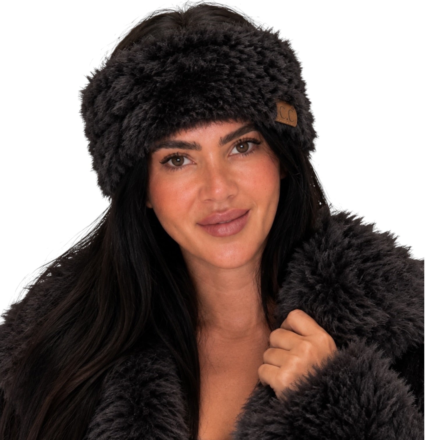 A woman wearing a dark brown faux fur headband with a small brown leather logo patch and a matching dark brown faux fur coat, set against a solid white background.