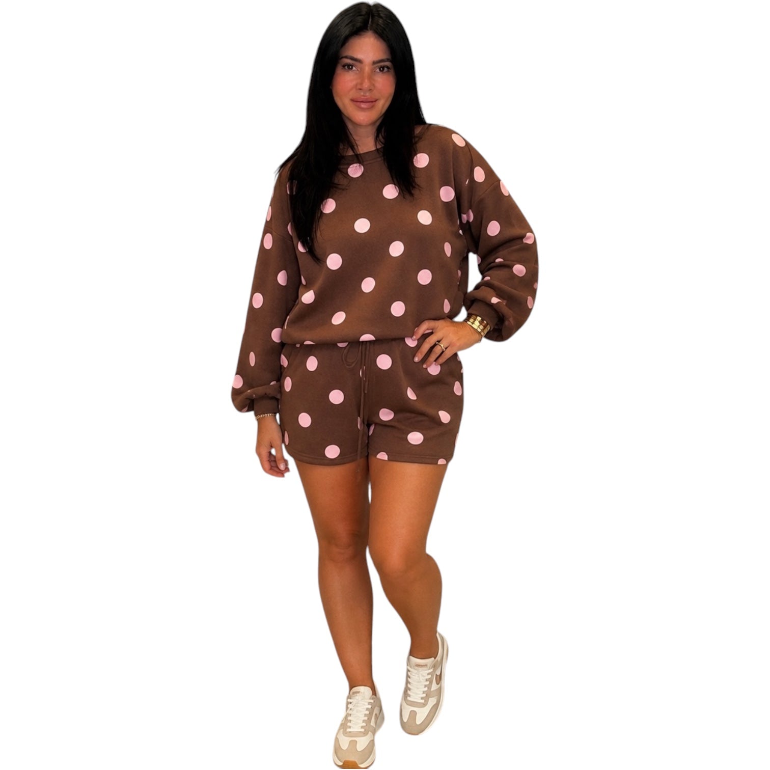 A woman modeling a brown two-piece lounge set with pink polka dots, consisting of a long-sleeve sweatshirt and matching shorts, paired with beige sneakers on a white background.