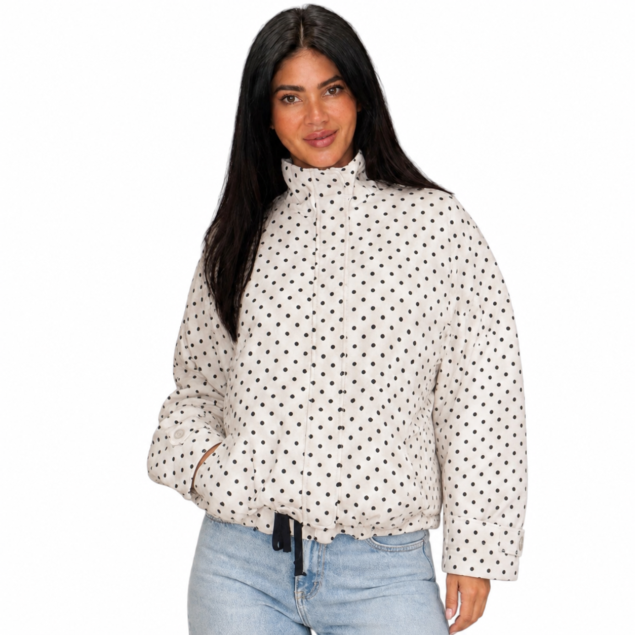 A woman modeling a cream-colored, high-neck, long-sleeve puffer jacket with a black polka dot pattern, paired with light-wash blue jeans against a plain white background.