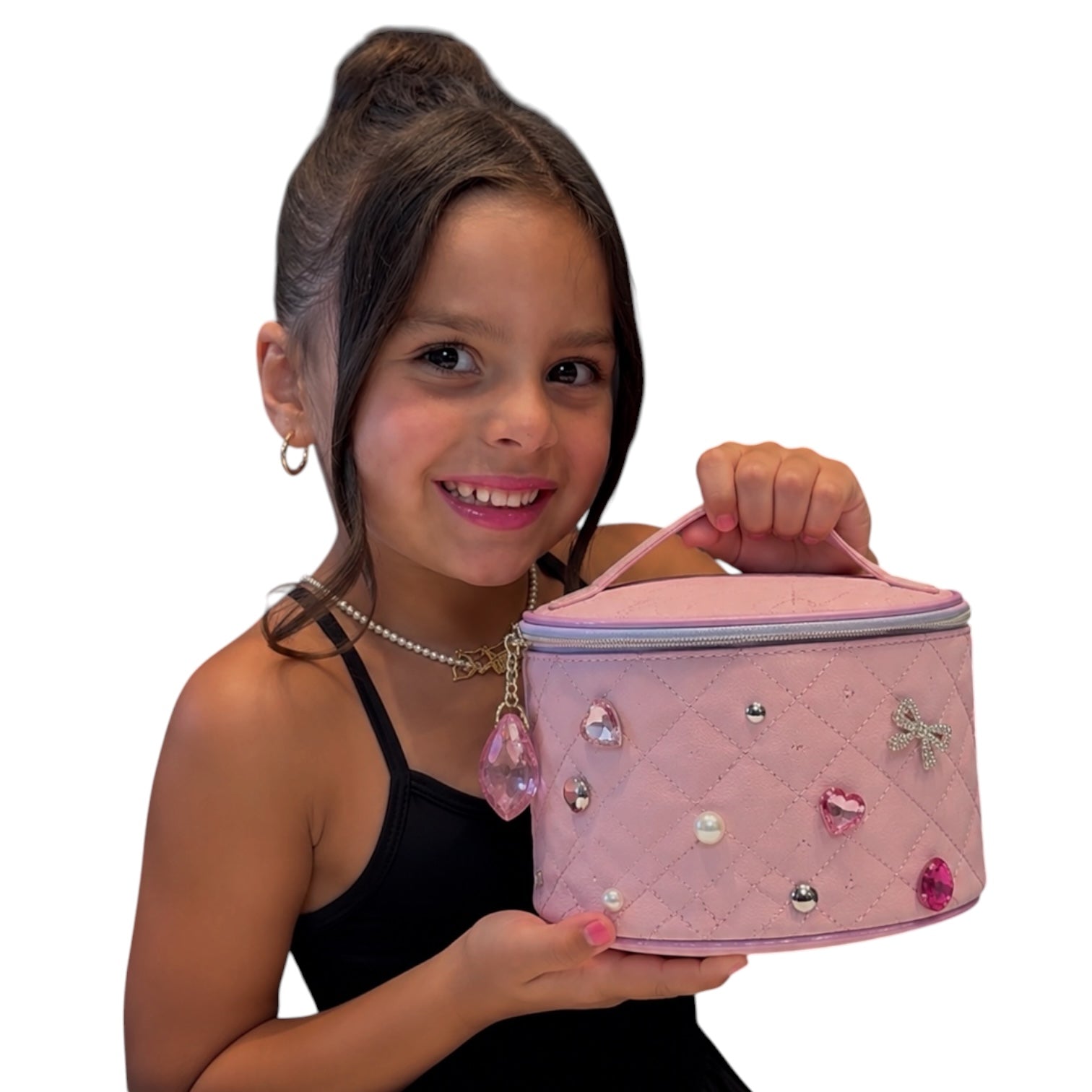 A young girl with her hair in a high bun holds a pink quilted cosmetic bag decorated with rhinestones and a dangling pink teardrop charm, against a plain white background.