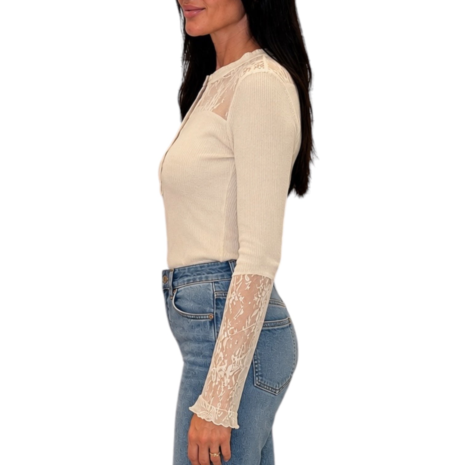 A woman shown in side profile wearing a fitted cream ribbed long-sleeve top with sheer lace panels on the shoulders and one sleeve, paired with blue jeans, against a plain white background.