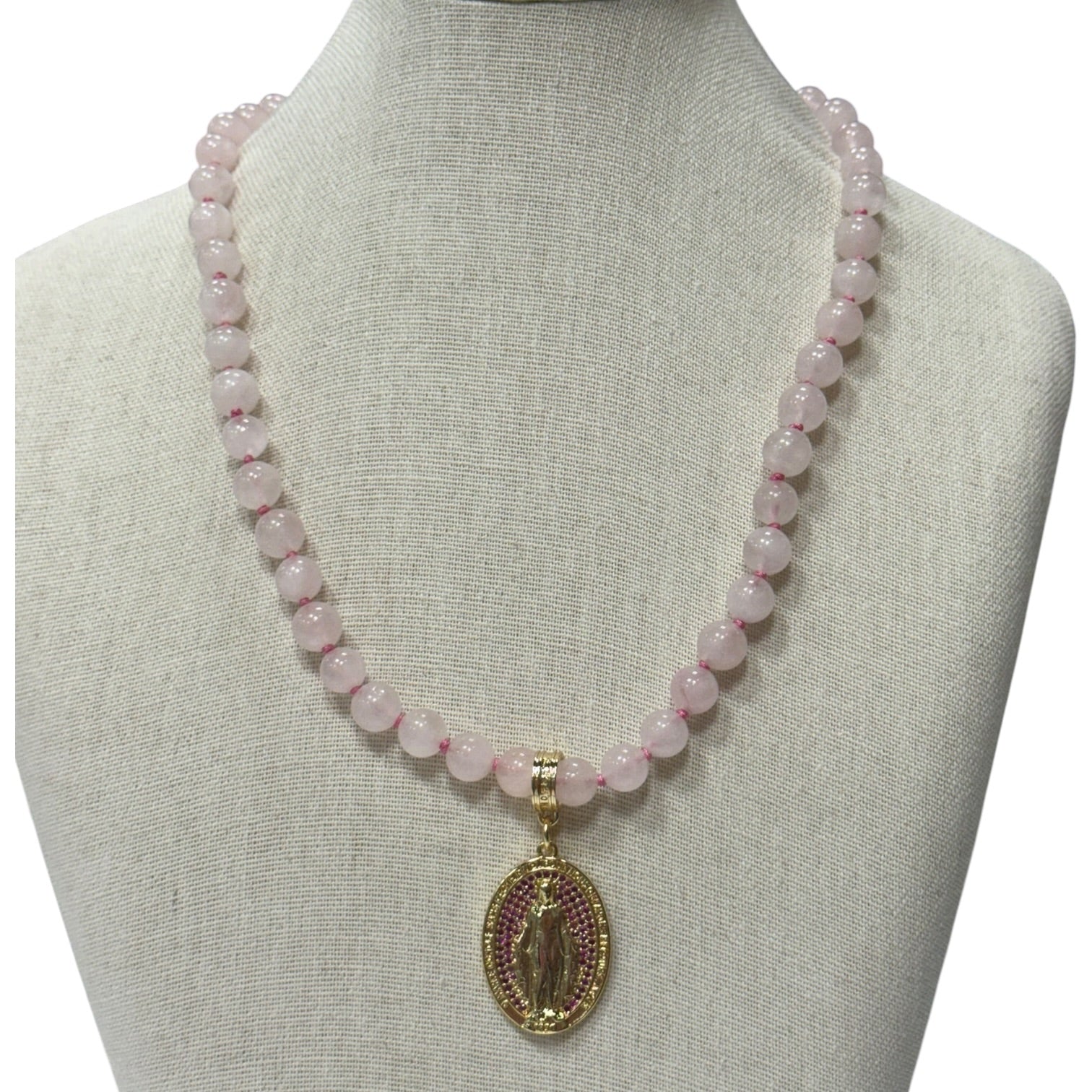 A pink stone bead necklace with a gold-toned oval religious pendant displayed on a beige fabric jewelry bust against a white background.