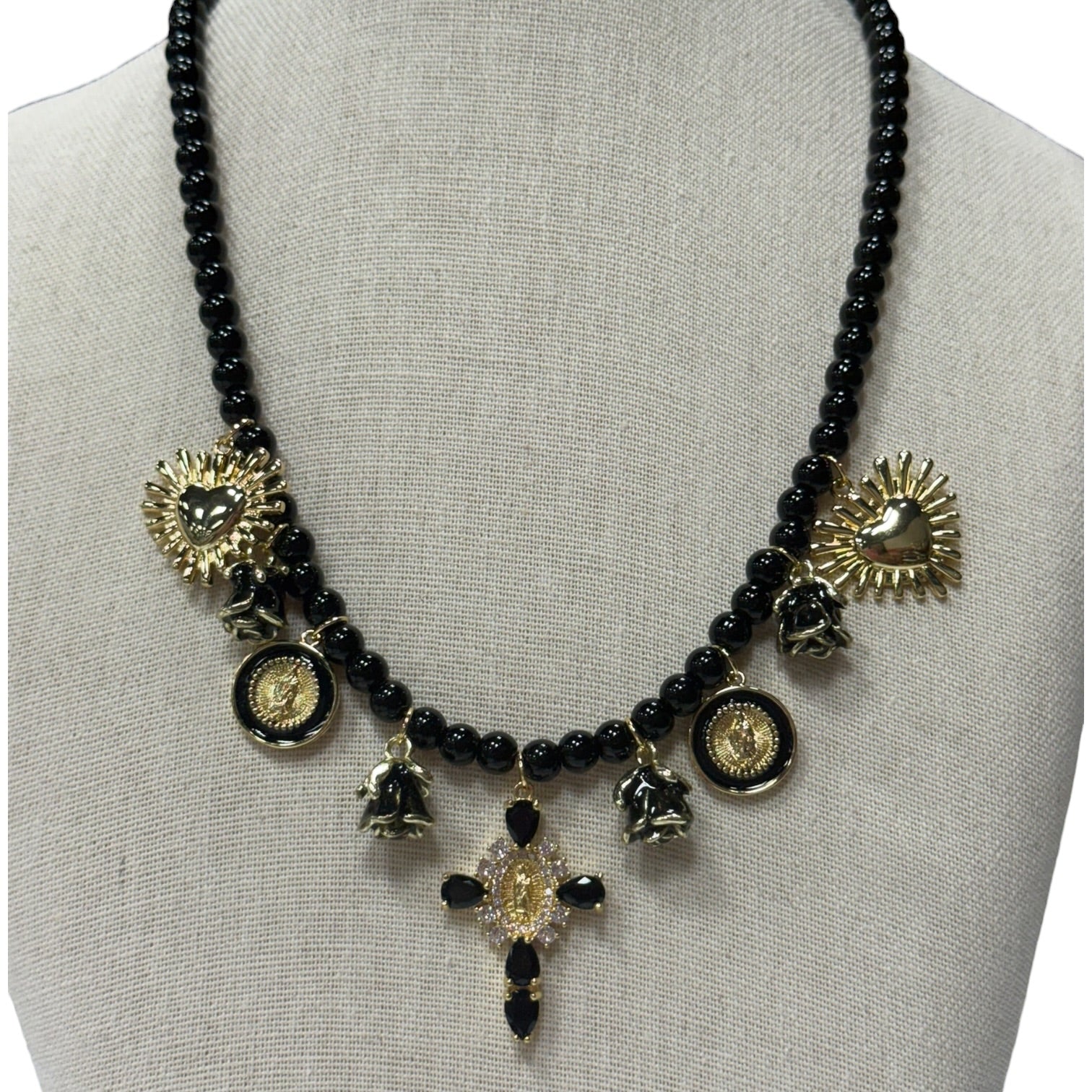 A black beaded statement necklace displayed on a neutral fabric mannequin bust. The necklace features multiple gold-toned charms, including sunburst hearts, circular medallions, small bells, and a central cross pendant embellished with gold, clear stones, and black beads.