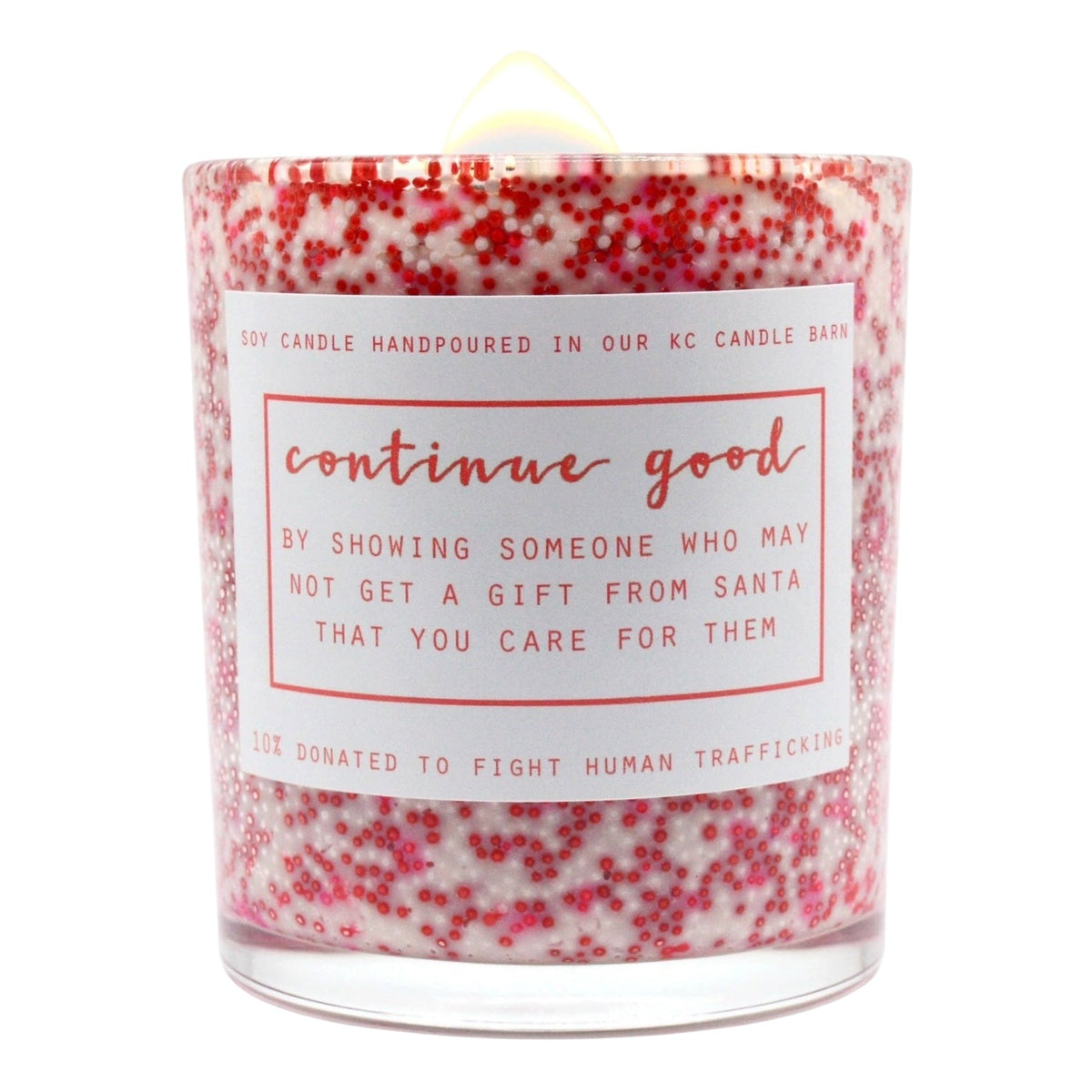 A hand-poured soy candle in a clear glass jar filled with white wax and dense pink and red glitter flakes. The jar has a white paper label with pink text that reads 'continue good' and a humorous message about showing someone who may not get a gift from Santa.