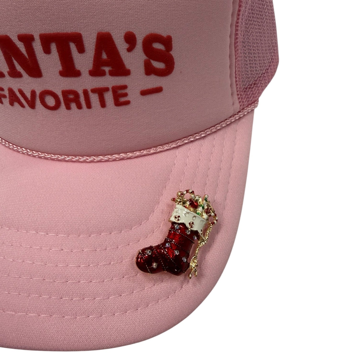 A close-up of a light pink trucker hat with red embroidered text reading 'SANTA'S FAVORITE' on the front panel and a small red-and-white stocking embellishment on the brim, shown against a white background.
