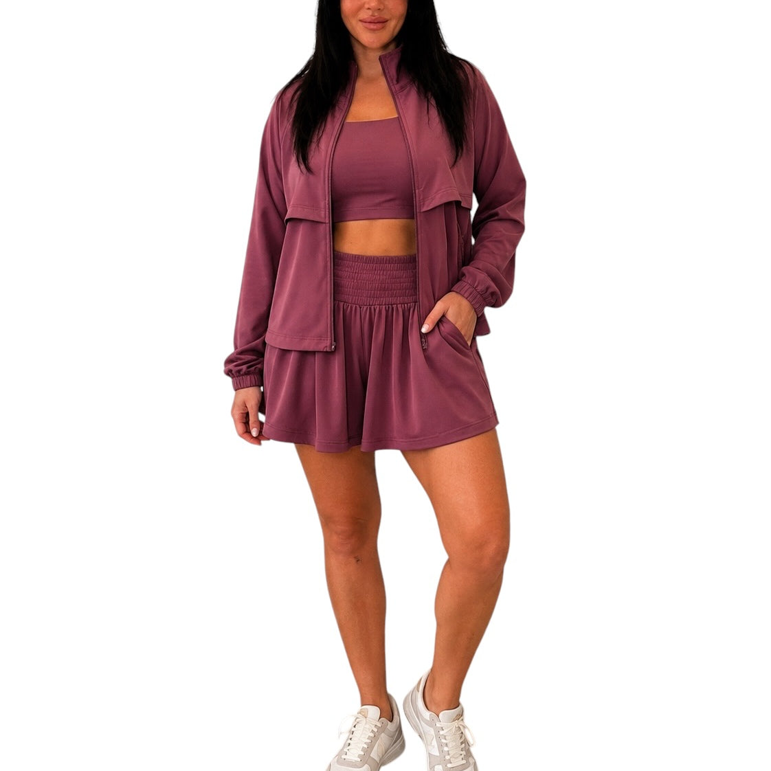 A woman modeling a matching mauve workout set consisting of a cropped tank top, high-waisted shorts, and a zip-up jacket, paired with white sneakers, against a plain white background.