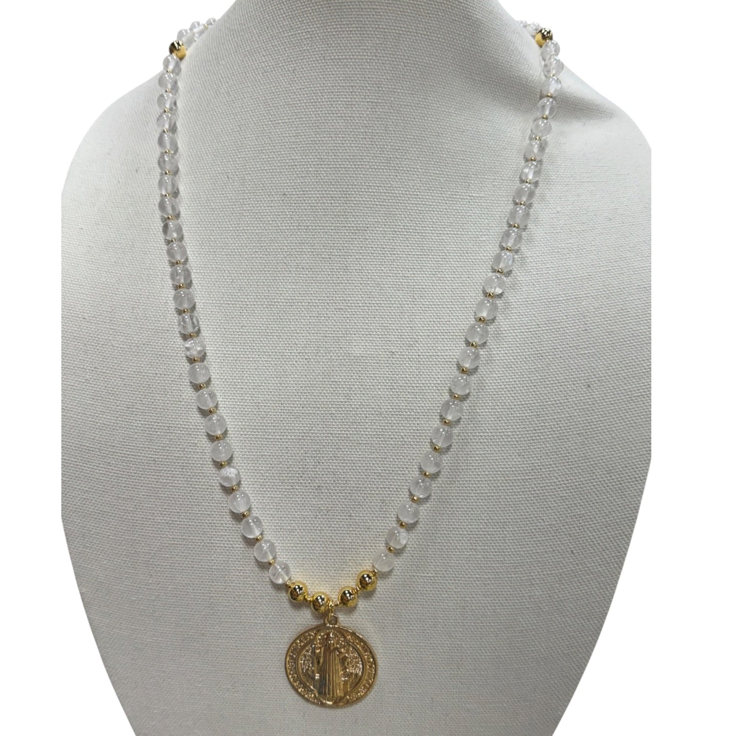 A beaded necklace displayed on a white fabric mannequin bust. The necklace features a strand of clear faceted beads separated by small gold spacer beads, with a gold Saint Benedict medallion pendant at the center.