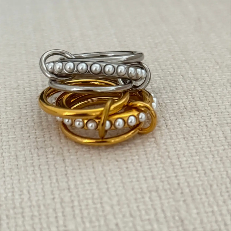 Two rings, one silver and one gold, with pearl accents on a beige fabric background.