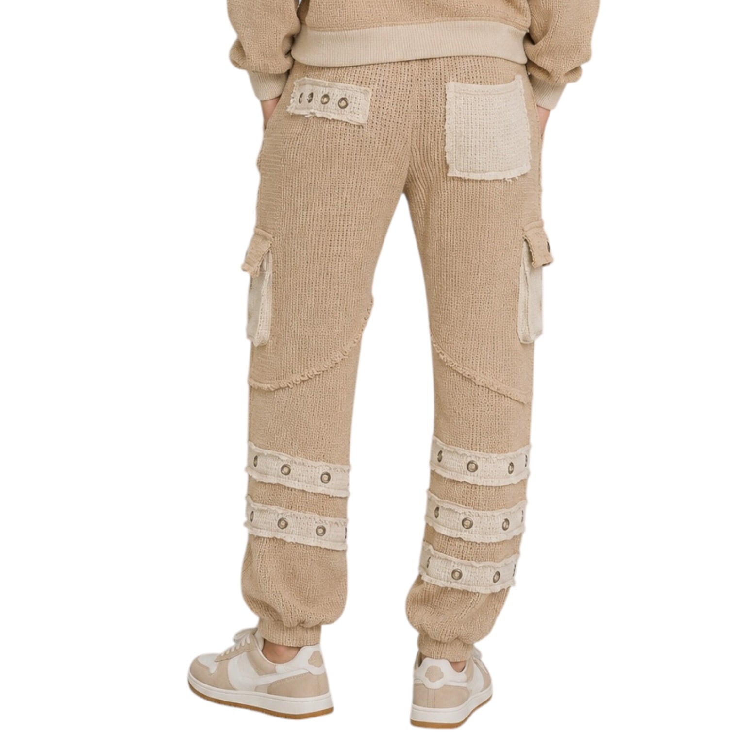 A person shown from the back wearing beige textured knit cargo pants with cream-colored patch pockets, decorative straps, and grommet details, paired with white sneakers against a plain white background.