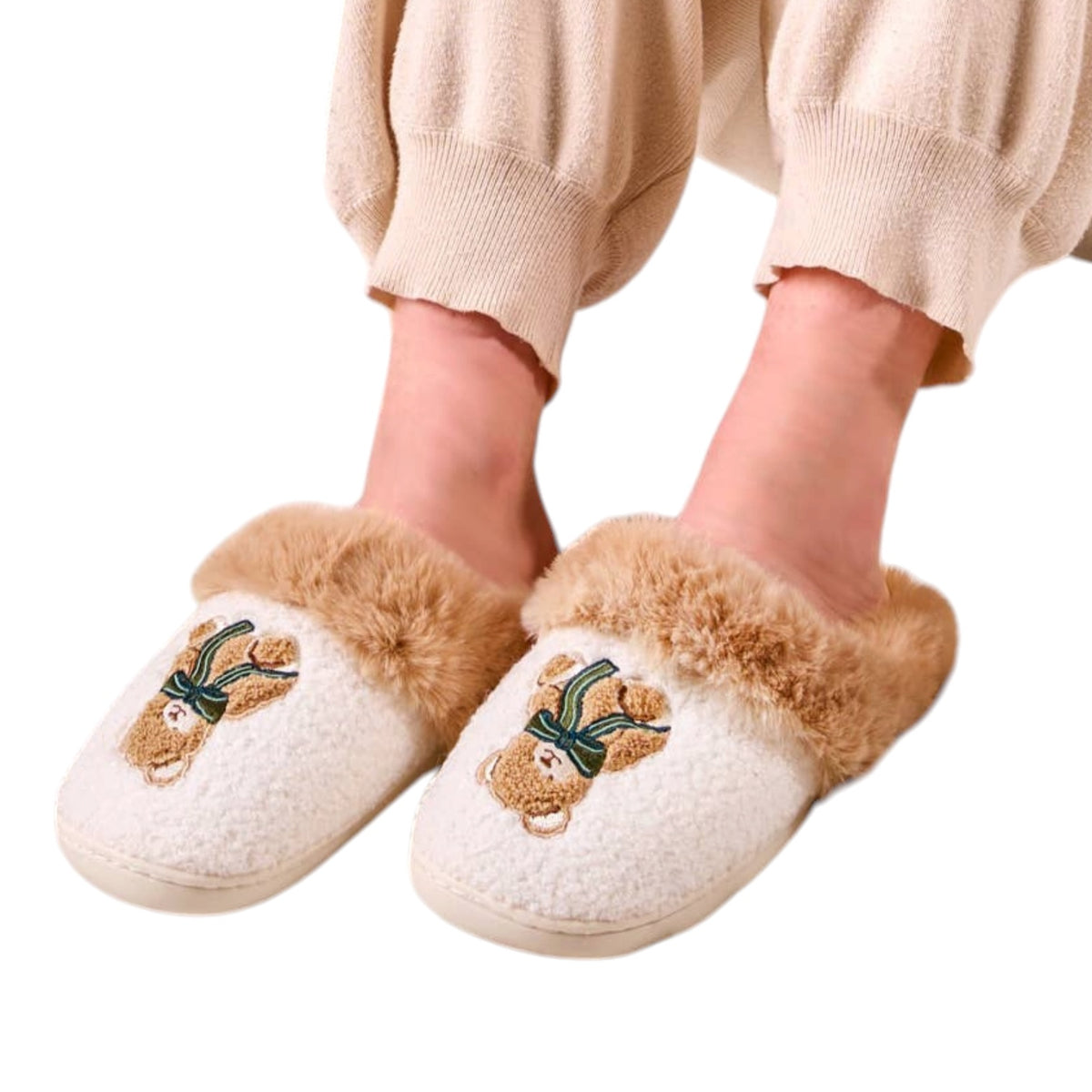 A person wearing fluffy cream-colored slip-on slippers with tan faux-fur trim and embroidered teddy bear faces with green bows, shown against a plain white background.