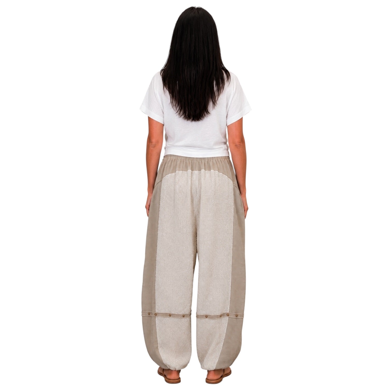 A woman viewed from behind wearing loose, wide-leg beige pants with a white t-shirt and brown sandals, set against a plain white background.