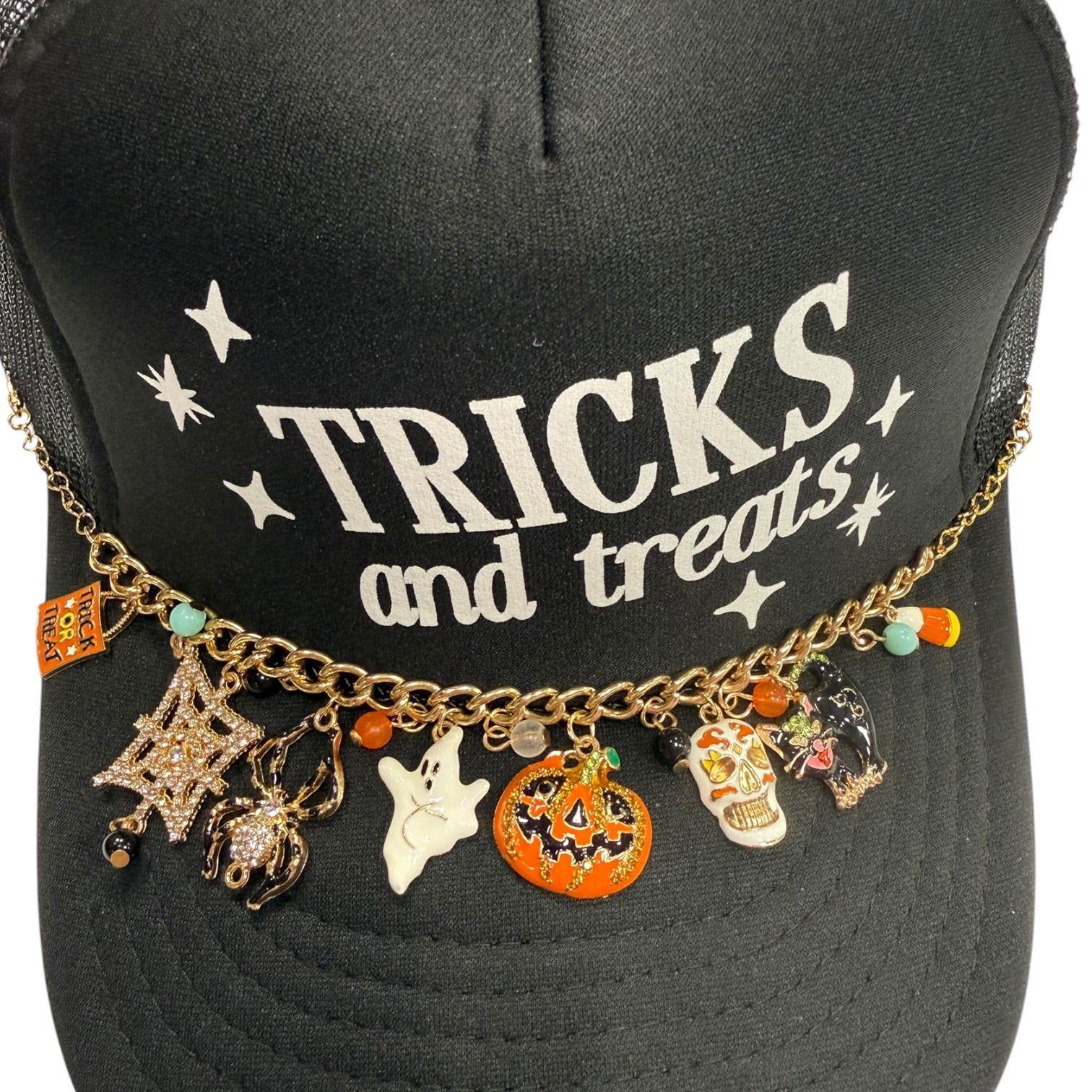 A black trucker hat with 'TRICKS and treats' printed in white on the front, decorated with a gold chain across the brim featuring various Halloween charms including a spider, ghost, pumpkin, and sugar skull.