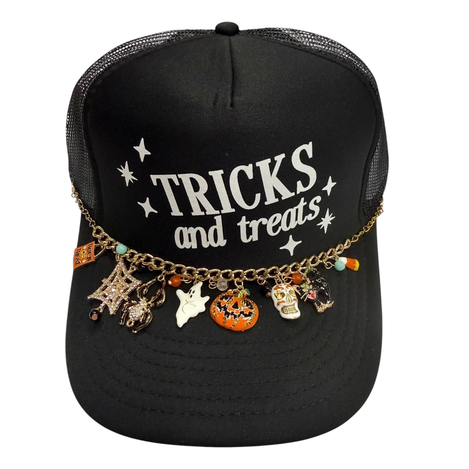 A black trucker hat with 'TRICKS and treats' printed on the front, decorated with a gold chain and various Halloween-themed charms including a pumpkin, ghost, skull, spider, and candy.