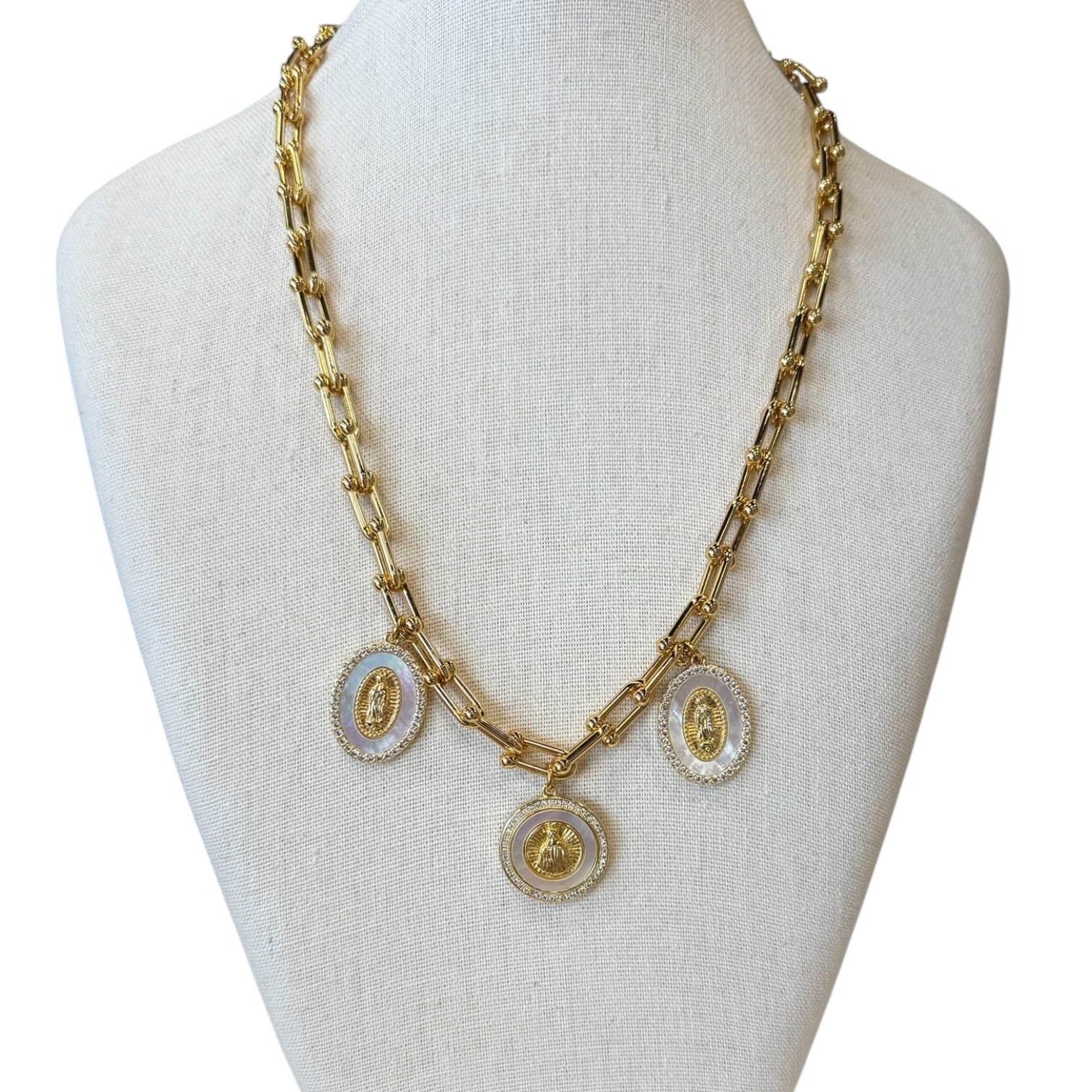 A gold-tone chain necklace displayed on a cream fabric mannequin bust. The necklace features three oval pendants with white inlay and gold religious-style portraits.