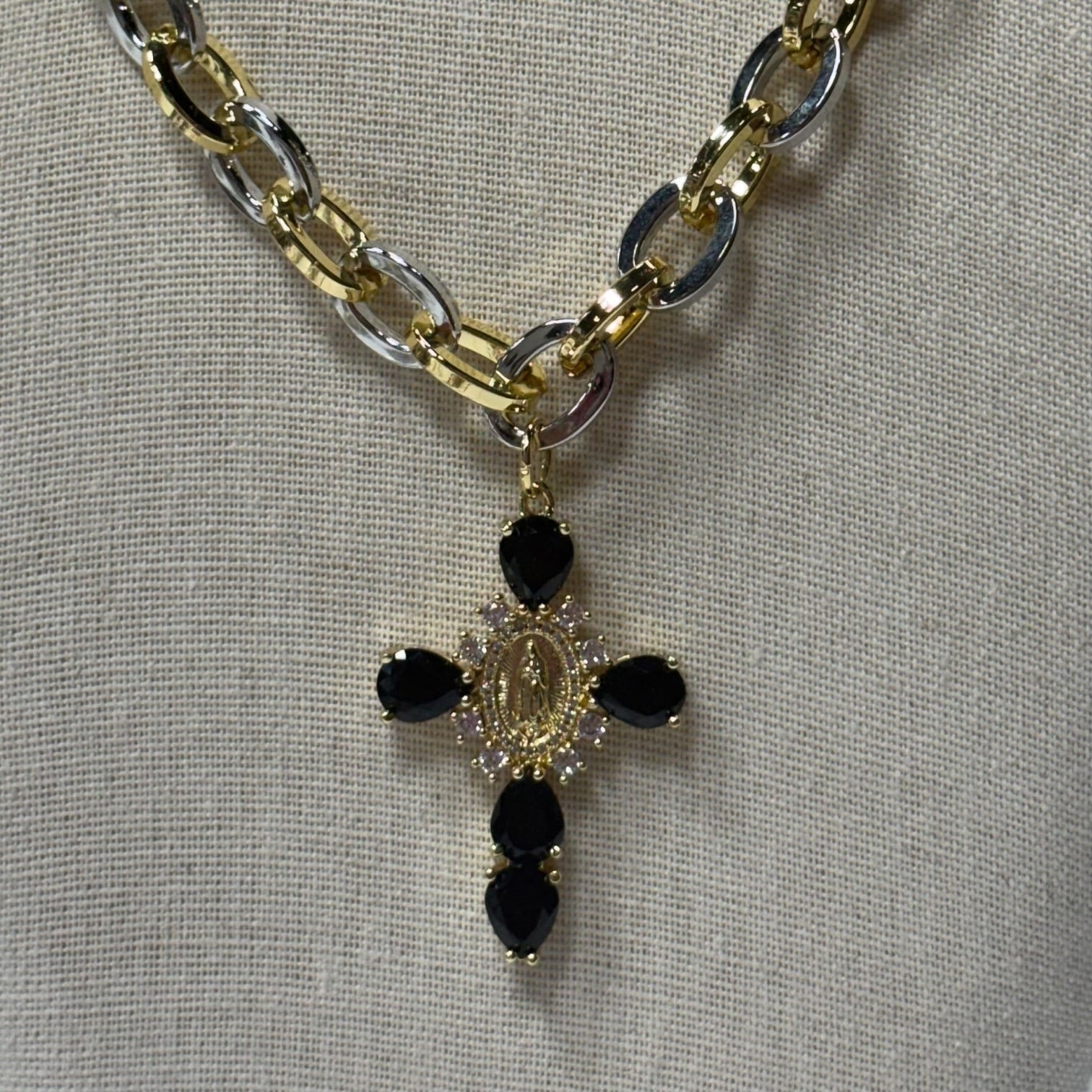 A gold and silver chain necklace with a cross pendant featuring black teardrop-shaped stones and a central gold-toned oval detail, displayed on a textured beige fabric background.
