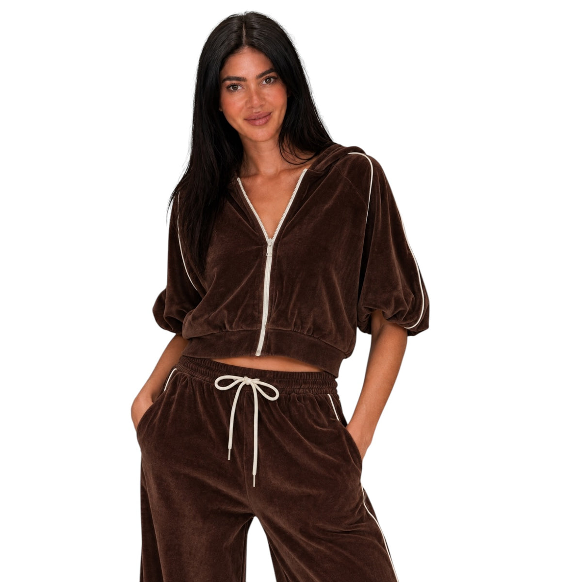 A woman modeling a brown velvet two-piece tracksuit with white piping and white drawstrings, consisting of a cropped zip-up hoodie and matching sweatpants, against a plain white background.