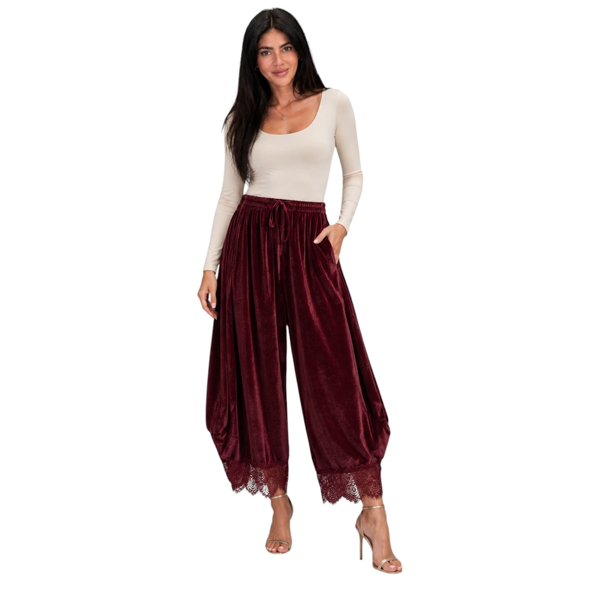 A woman modeling a pair of high-waisted, balloon-style harem pants in deep burgundy velvet with lace trim at the hems, paired with a cream long-sleeve top and nude high heels against a plain white background.