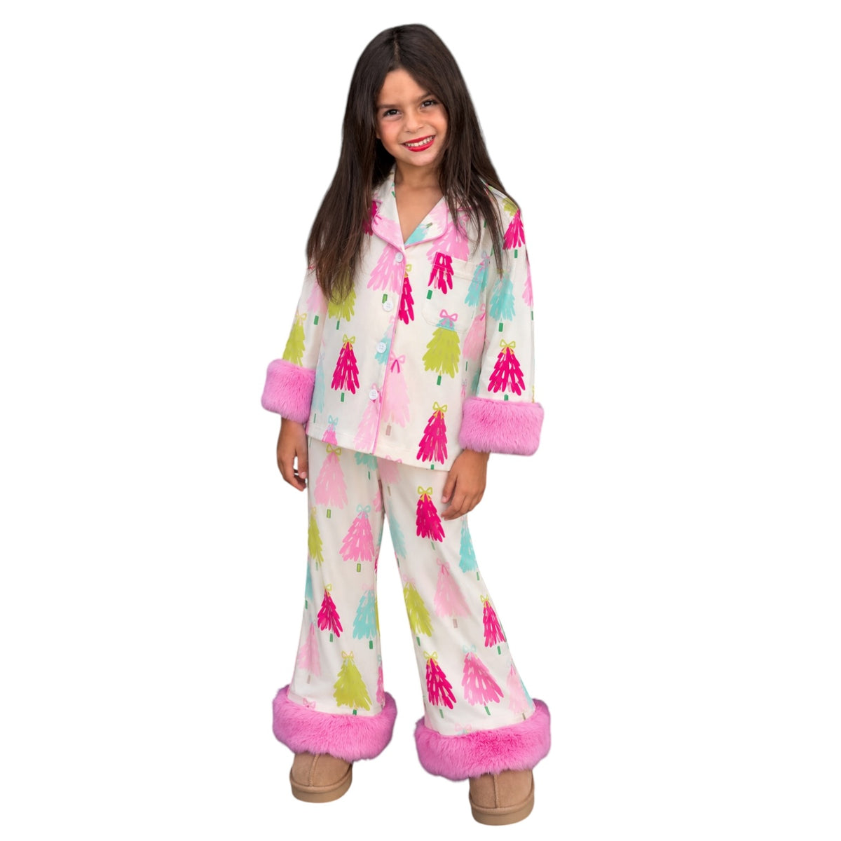 A young girl modeling a two-piece pajama set featuring a white base printed with colorful Christmas trees in pink, green, and blue, finished with bright pink faux-fur trim on the collar, cuffs, and hems.