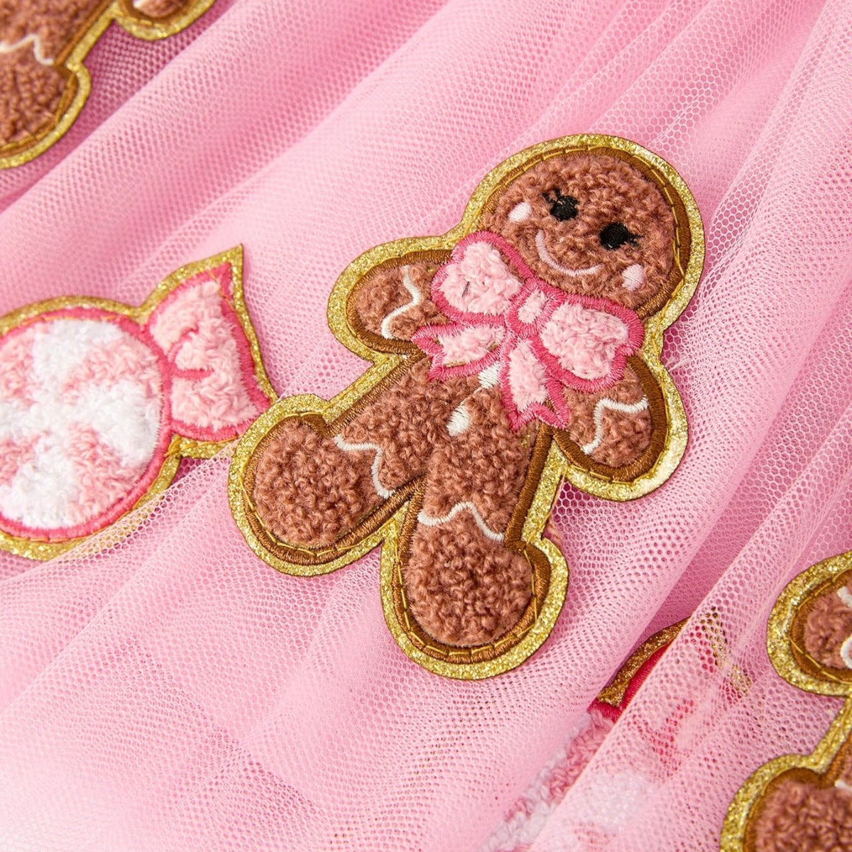 Close-up of pink tulle fabric featuring embroidered gingerbread cookies with gold glitter trim and pink bows, alongside a pink-and-white candy motif.