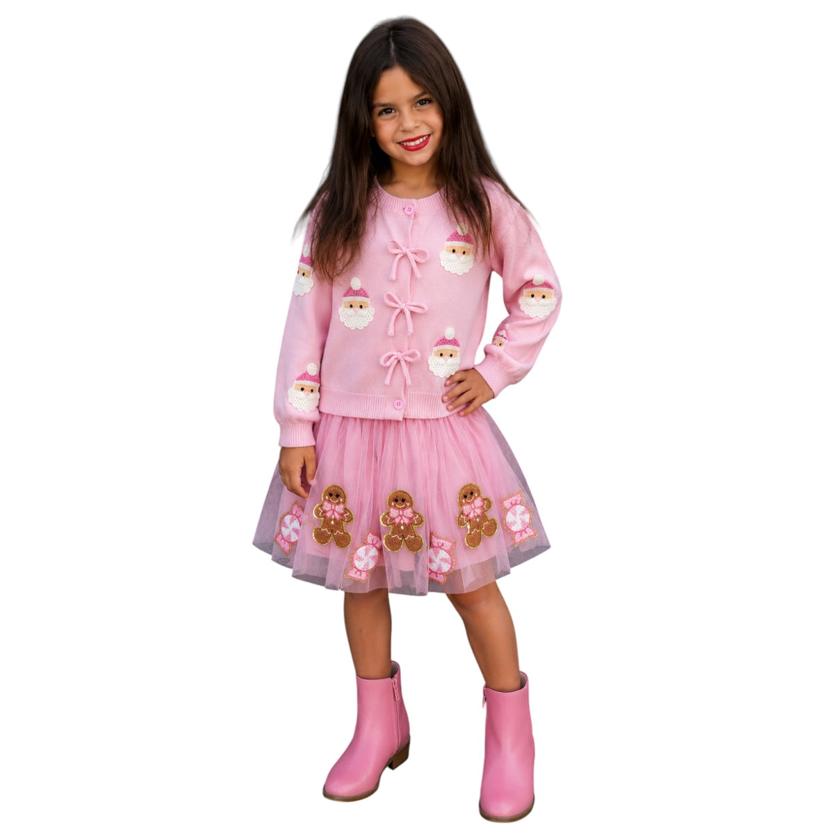 A young girl modeling a pink holiday-themed knit sweater dress with Santa Claus and gingerbread man motifs, paired with pink boots, on a solid white background.