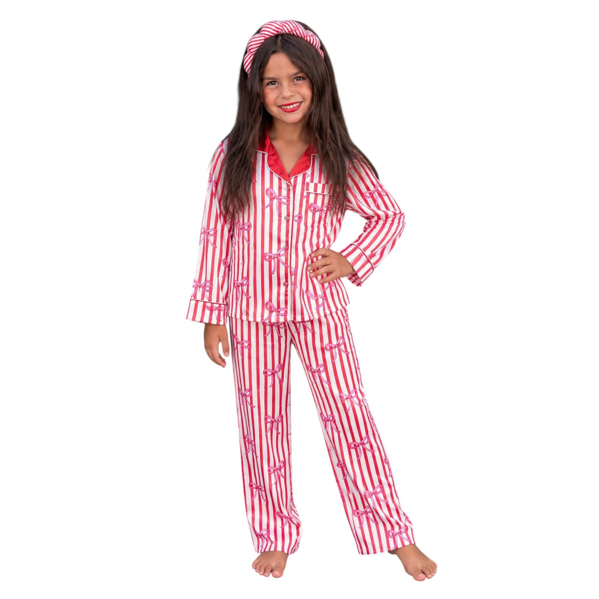 A young girl modeling a two-piece pajama set with red and white vertical stripes and pink bows, including a long-sleeve top and matching pants, along with a matching headband, on a solid white background.