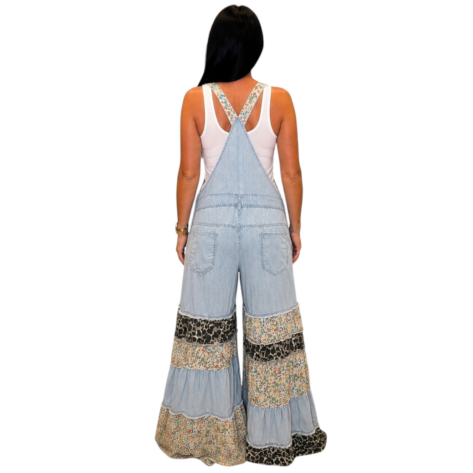 A woman shown from the back wearing a light blue denim overall jumpsuit with wide legs and mixed patterned ruffle hems, over a white tank top, against a plain white background.