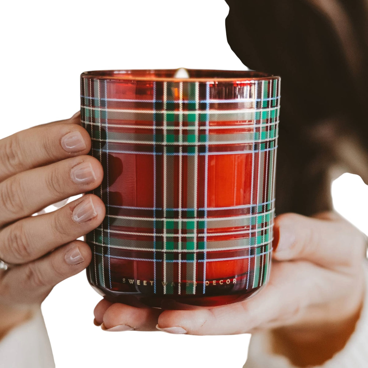 A close-up of a hand holding a red glass candle with a green, white, and blue plaid pattern. The brand name 'SWEET TREATS & DECOR' is embossed at the bottom of the glass.