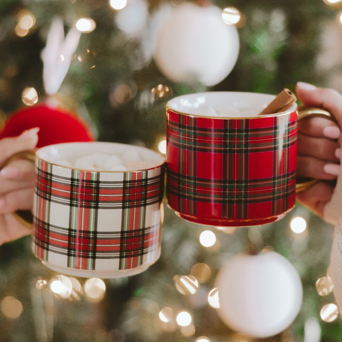 Two plaid mugs held in front of a decorated Christmas tree.