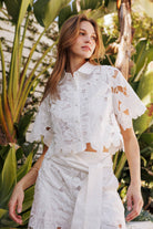 Model wearing a floral lace belted pants set, showcasing intricate lace details and a chic silhouette in a tropical setting.