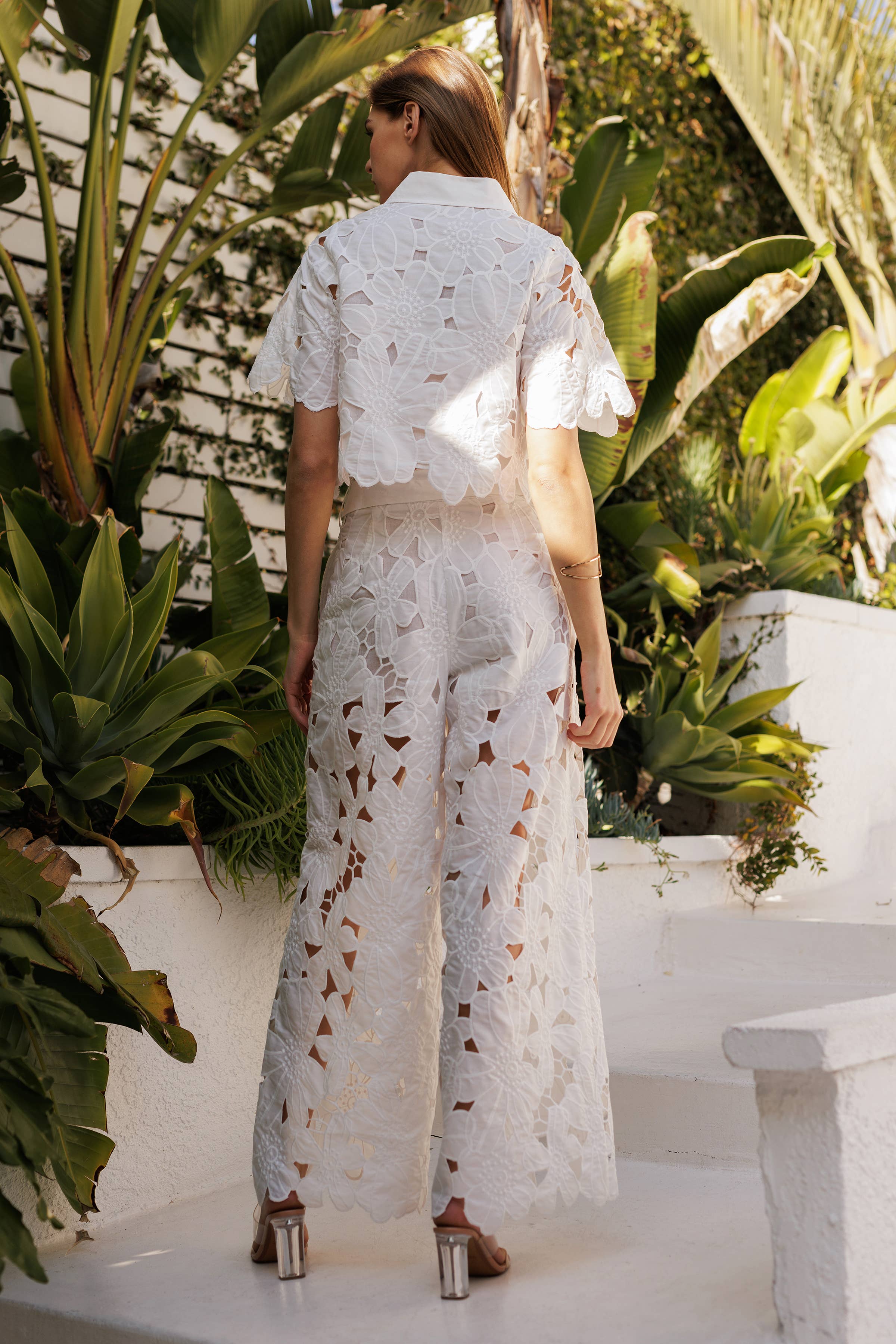 Back view of a woman in a floral lace belted pants set, surrounded by tropical greenery.