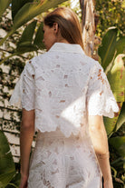 Back view of a woman wearing a white floral lace blouse among lush greenery, showcasing intricate lace details.